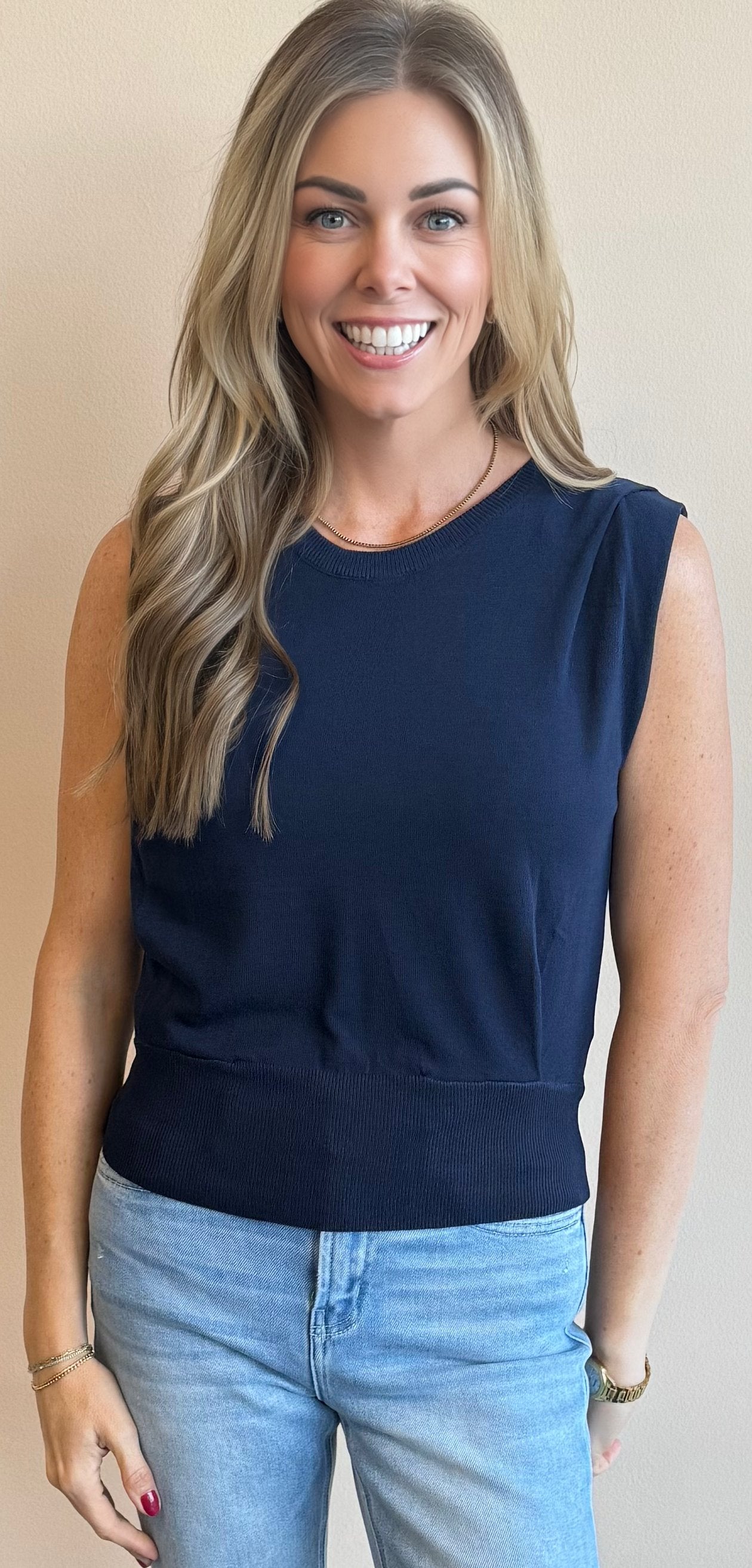 Woman wearing a navy sleeveless vest top and light blue jeans against a beige background