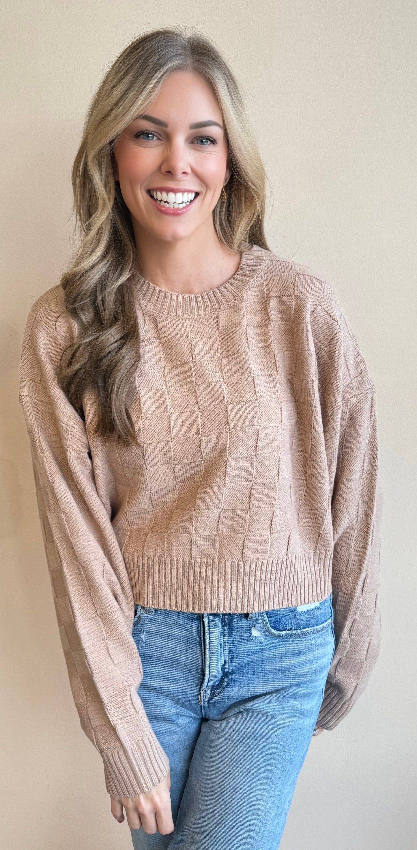 Woman wearing a checkboard pattern beige cropped sweater and blue jeans against a plain background