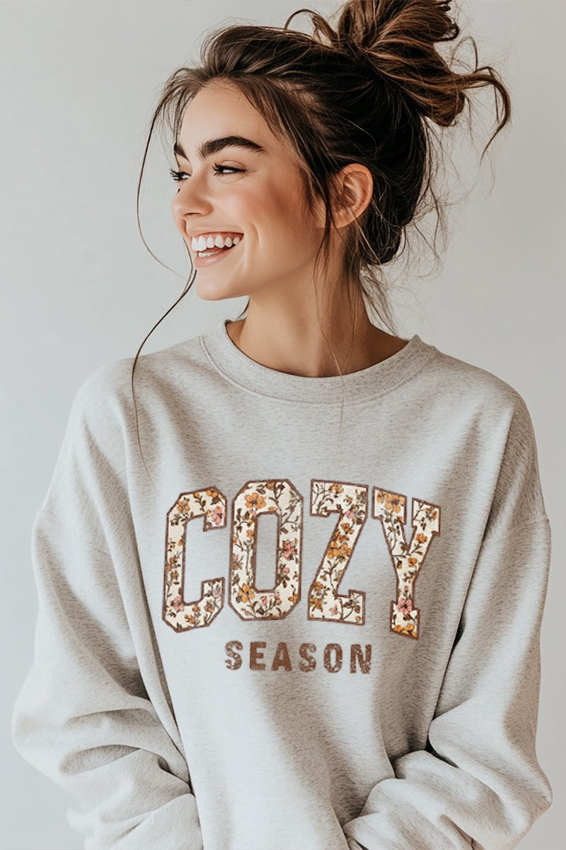 Woman wearing a 'Cozy Season' sweatshirt with floral letters on a plain background