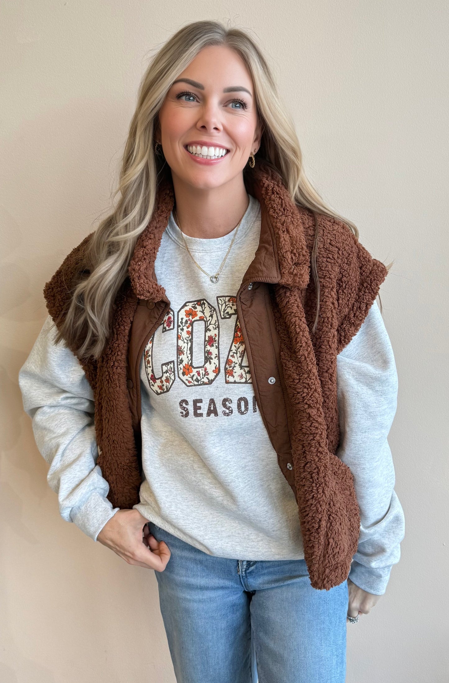 Woman wearing a brown fleece vest over a gray sweatshirt with floral text, standing against a beige wall.