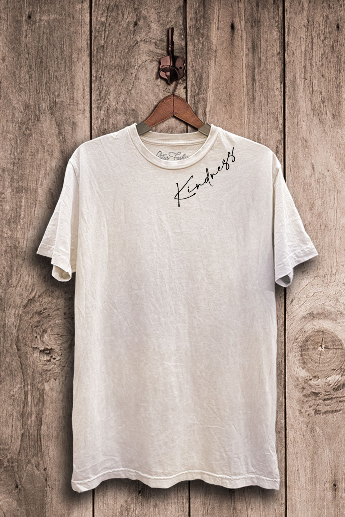 Cream, distressed t-shirt with 'Kindness' printed on it hanging on a wooden wall.