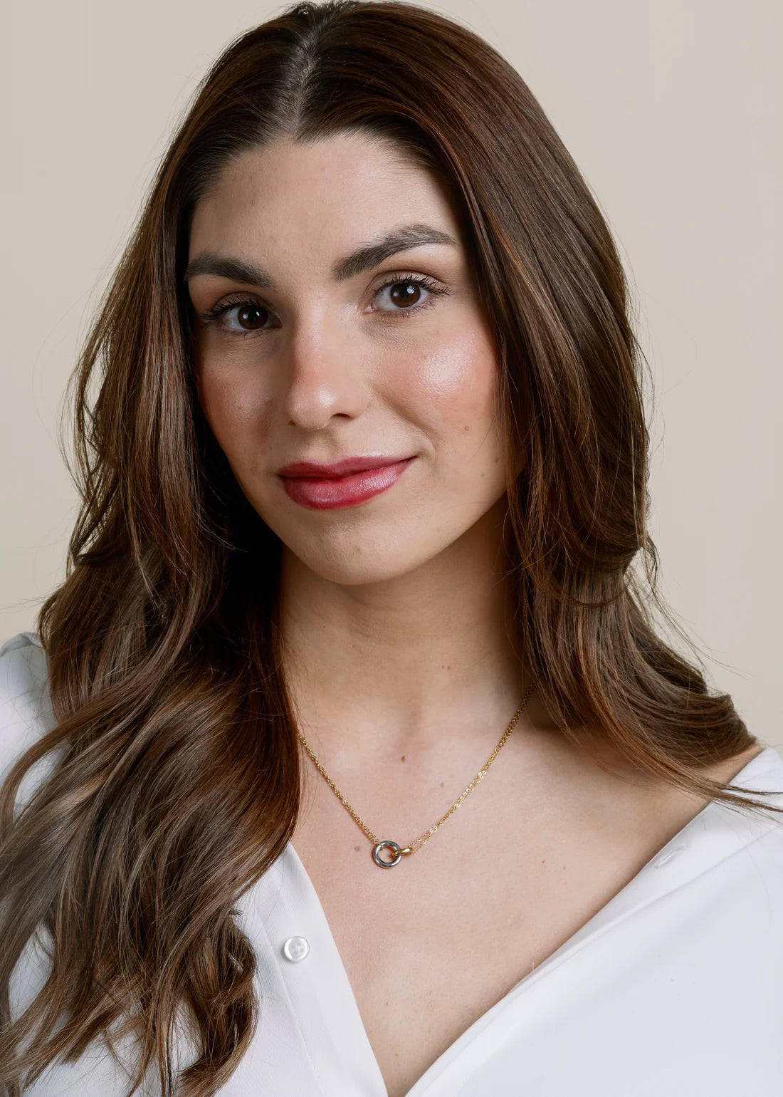 Woman with long brown hair wearing a white shirt and mixed-metal necklace with hooked circles pendant against a beige background