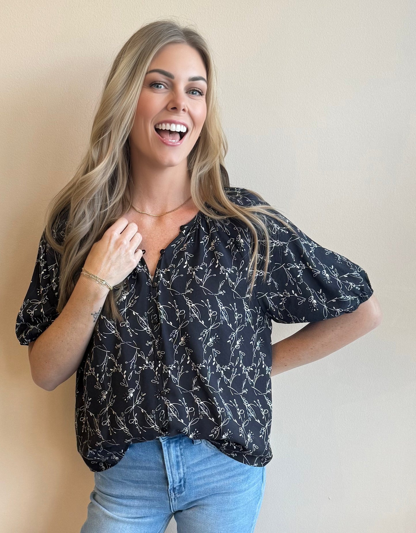 Woman smiling at the camera wearing a black floral patterned blouse and blue jeans against a beige background