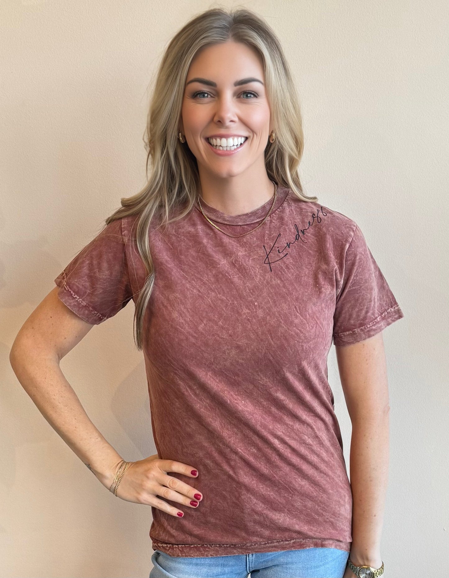 Woman smiling at the camera wearing a distressed maroon t-shirt with the word "Kindness" in a script font screen printed on the left should of the shirt.