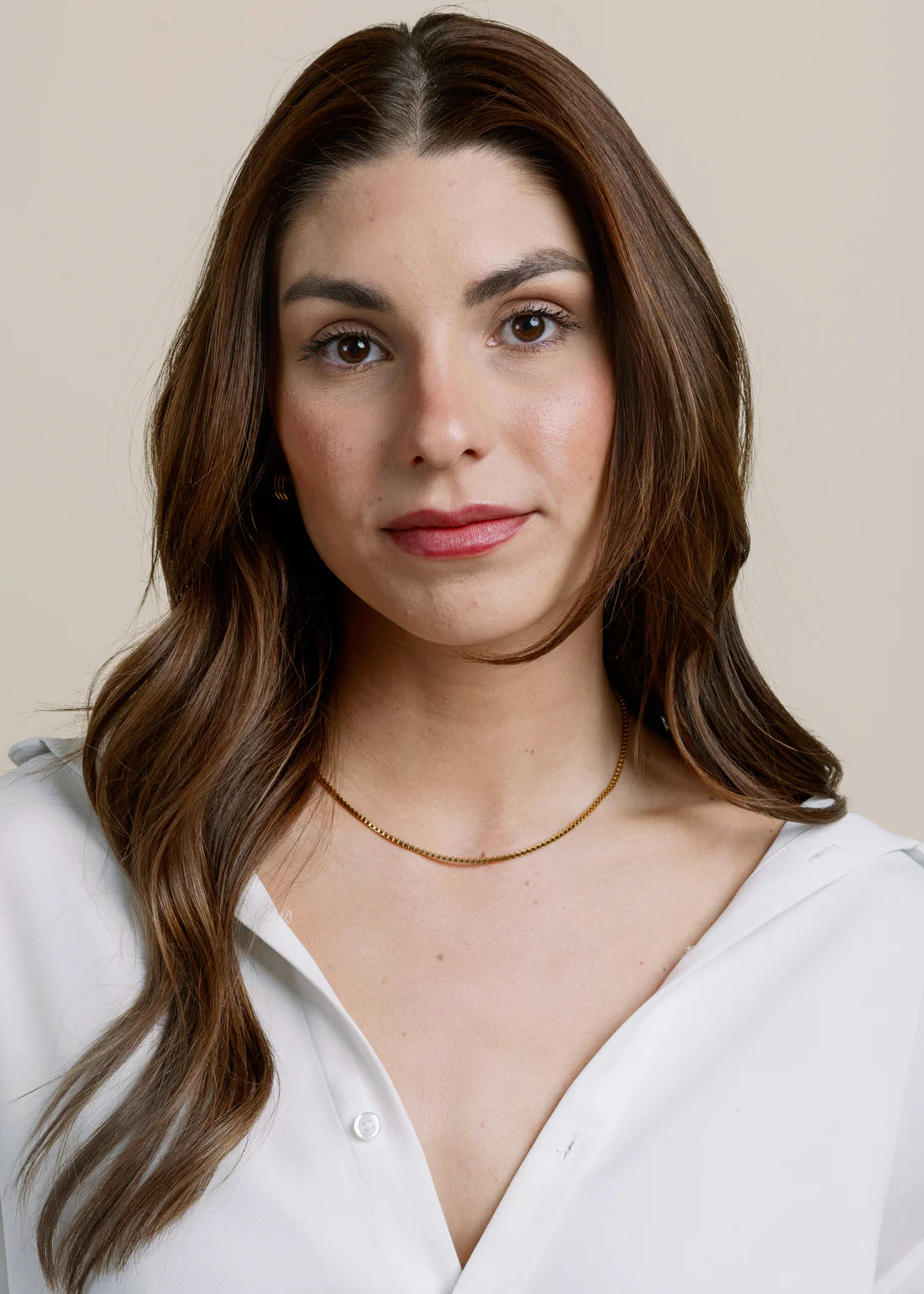 Woman with long brown hair wearing a white shirt and gold necklace against a beige background