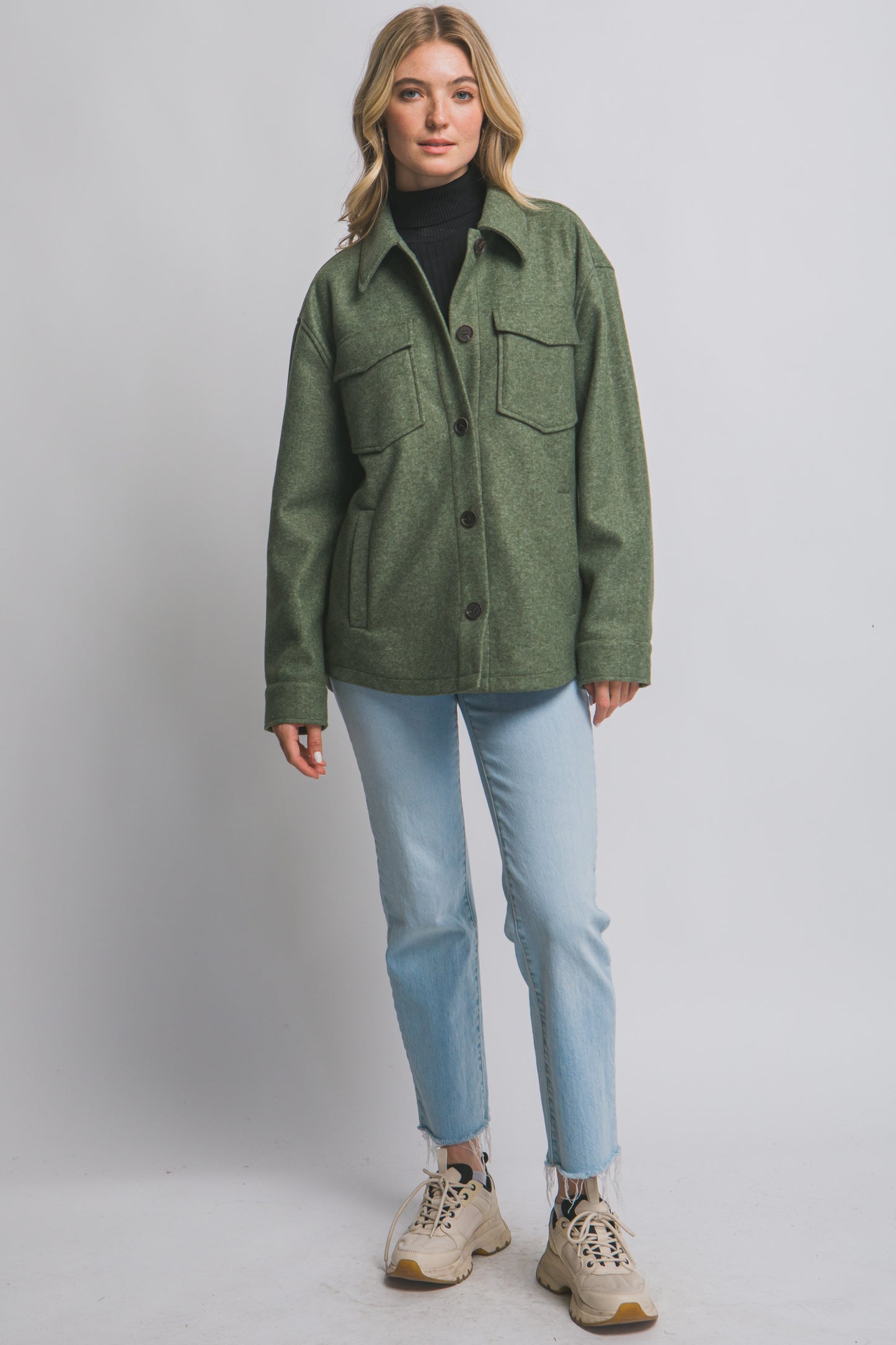 Woman wearing an olive green jacket, black turtleneck, light blue jeans, and beige sneakers on a gray background