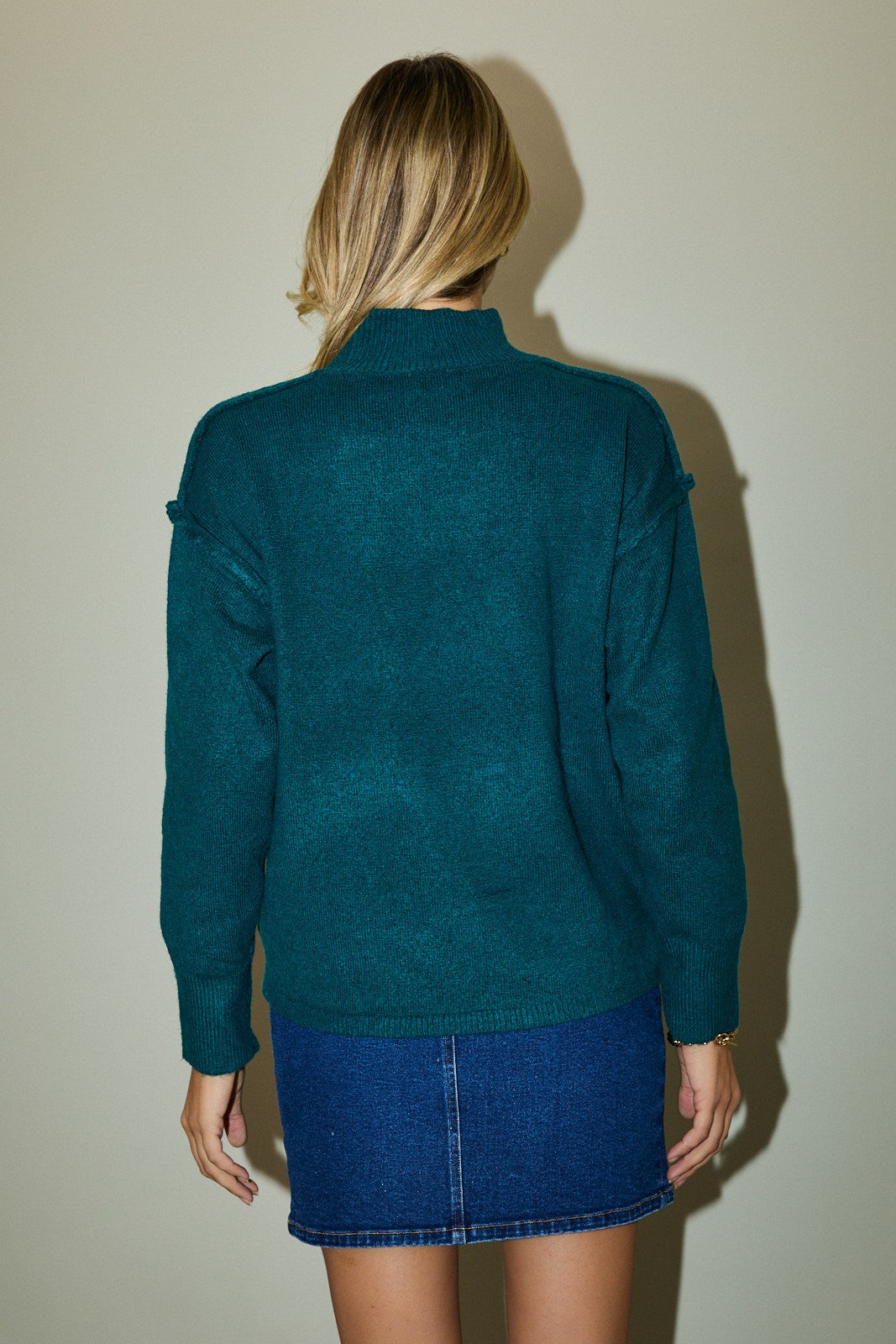 Person wearing a teal sweater and blue skirt against a plain background