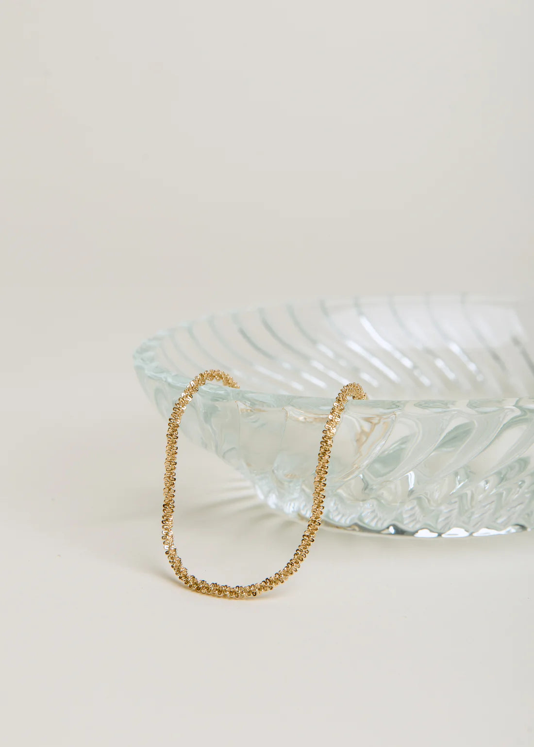 Clear glass bowl with gold chain bracelet on a light beige background