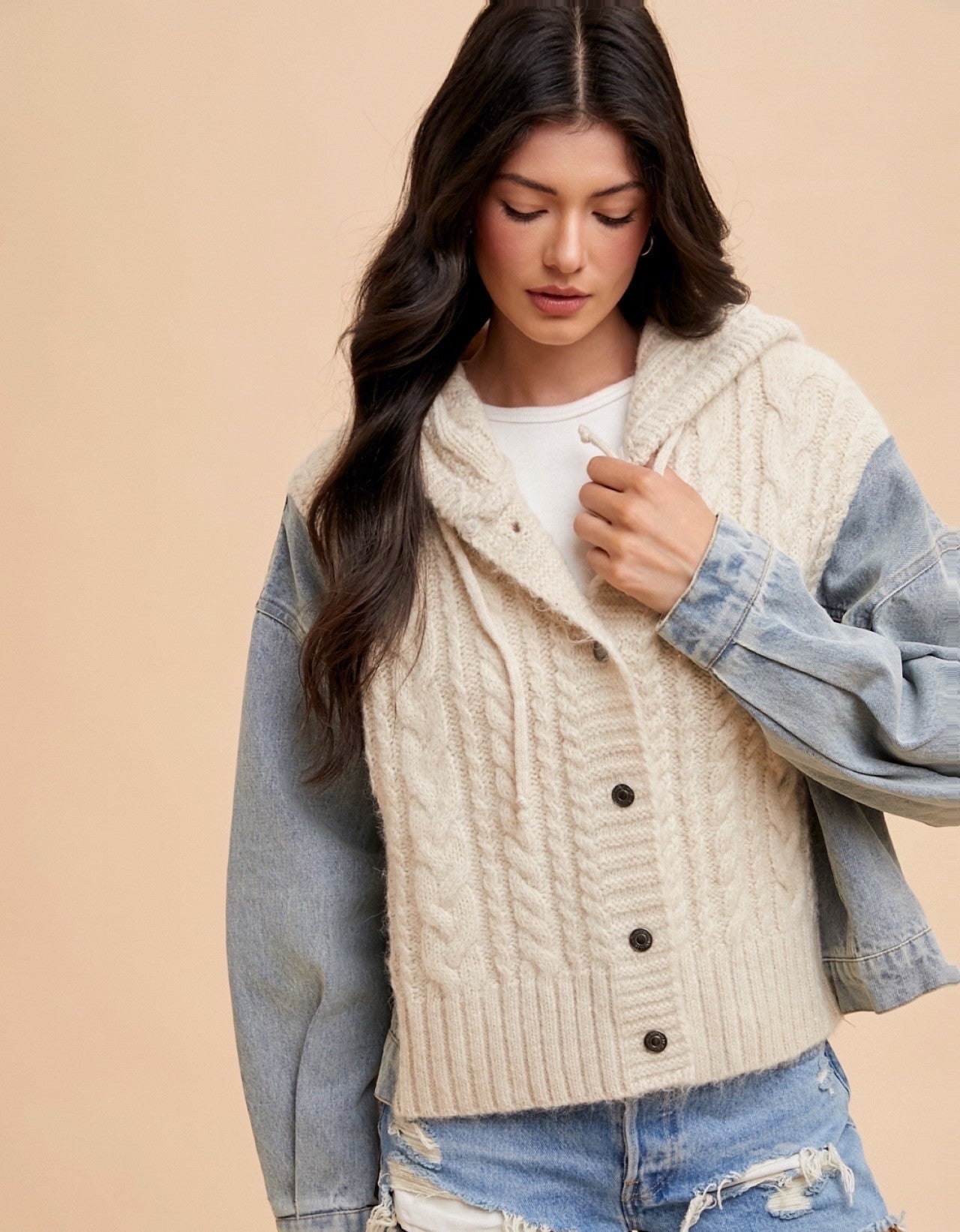 Woman wearing a cream cardigan, denim jacket, and shorts against a beige background