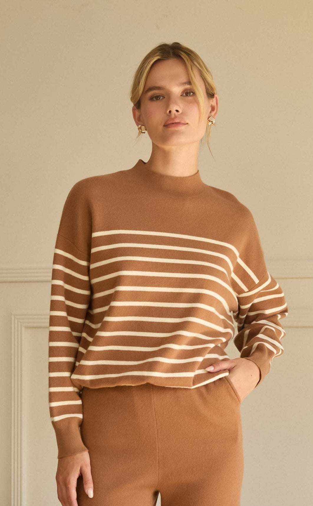 Woman wearing a brown and beige striped sweater and pants in a room with beige walls and a beige armchair.