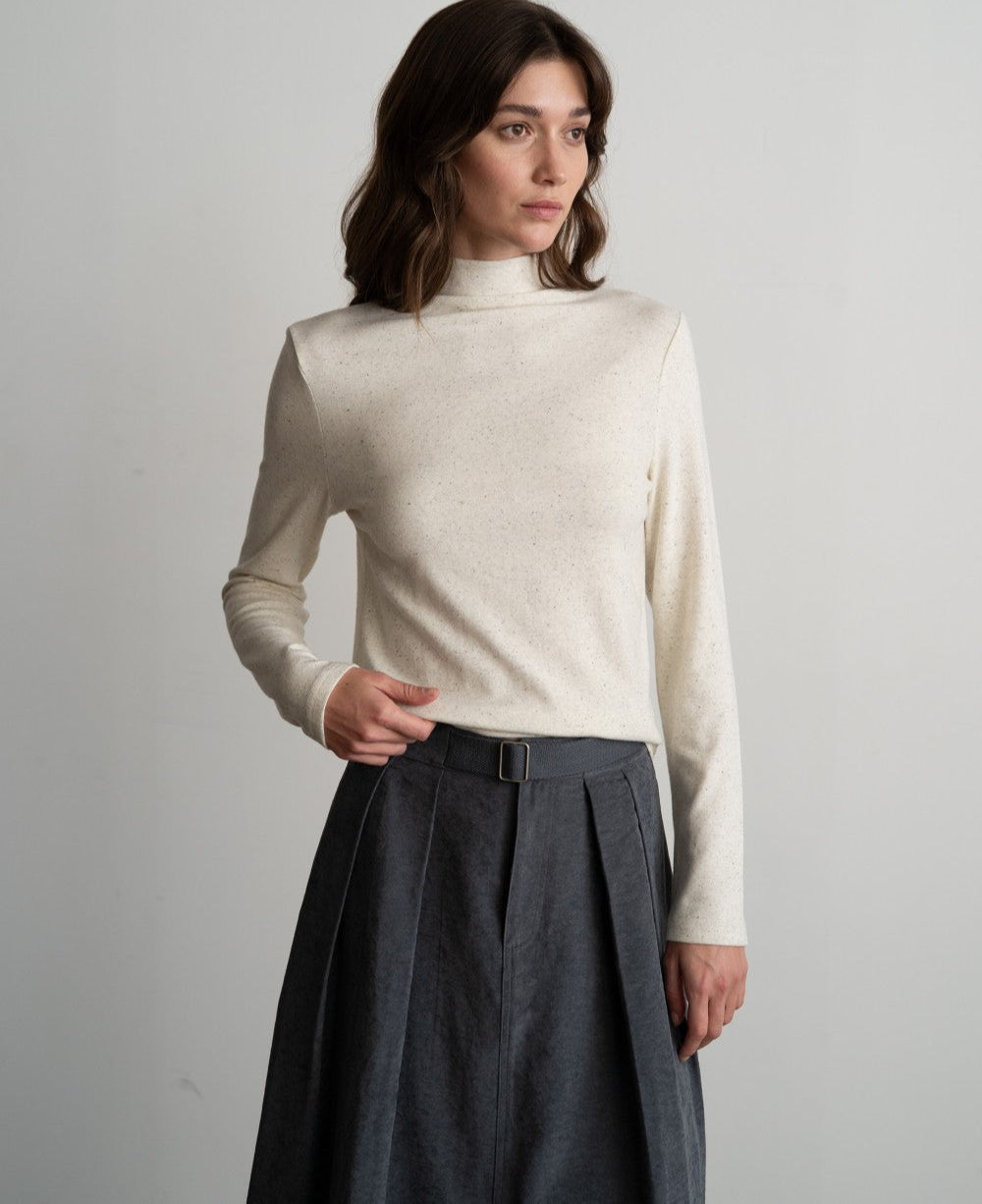 Woman wearing a white turtleneck sweater and dark gray pleated pants against a plain background