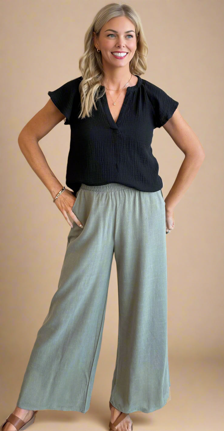 Woman wearing a black top and light green pants against a beige wall.