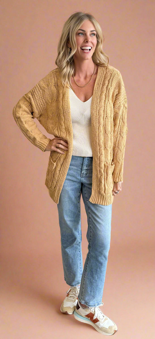 Woman wearing a beige cardigan, white top, and blue jeans against a beige wall.