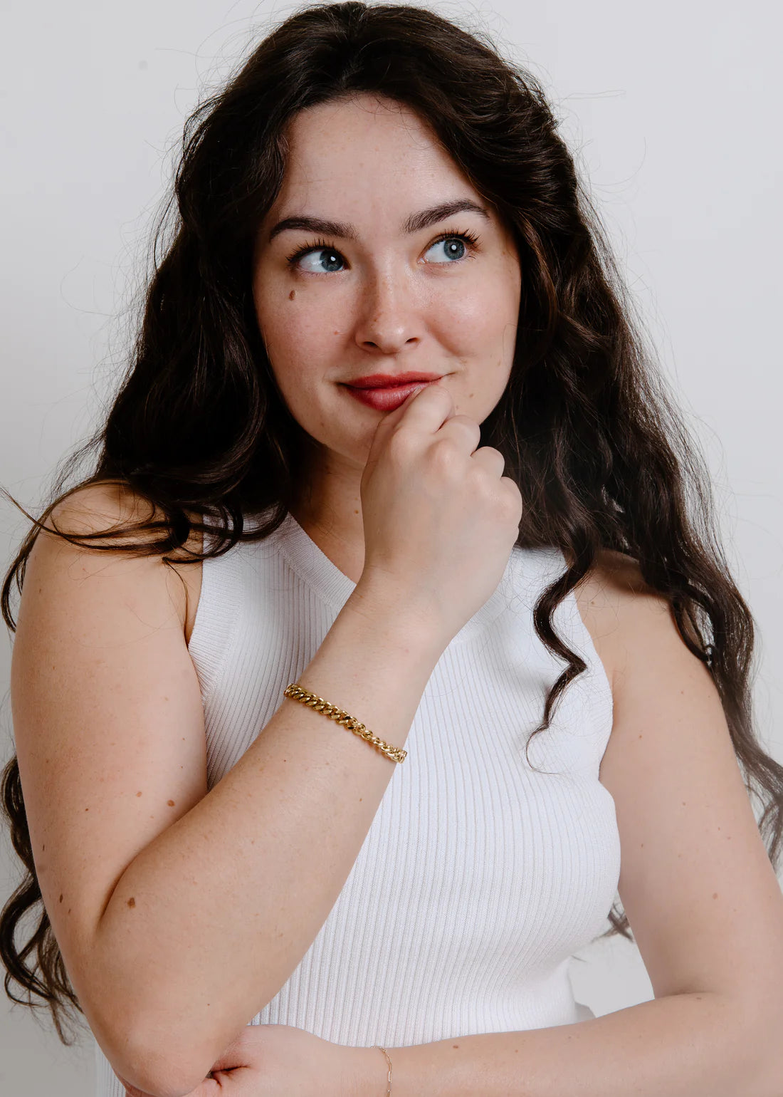 Woman with long dark hair wearing a white sleeveless top against a plain background her arm tilted up to her chin sporting a gold bracelet
