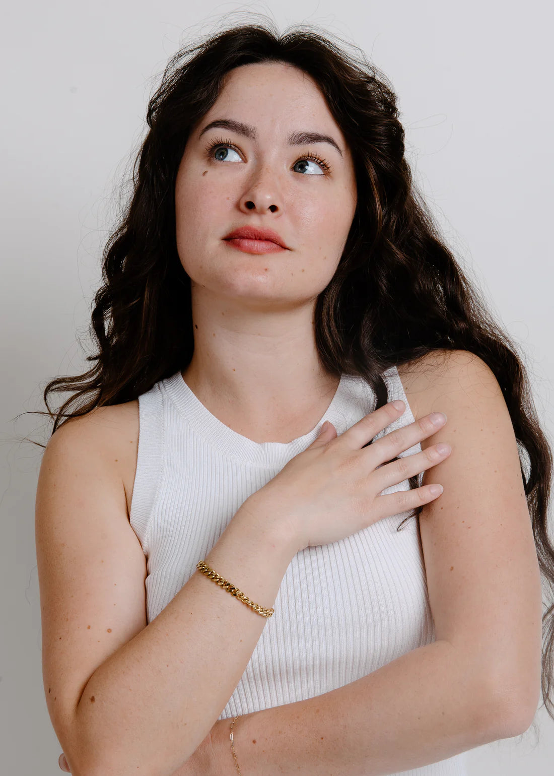 Woman looking up to the left holding her arm across her chest with a gold chain around her wrist