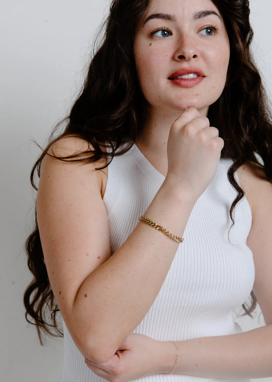 Woman wearing a white sleeveless top with a plain background holding her arm  vertical touching her chin with a gold bracelet around her write