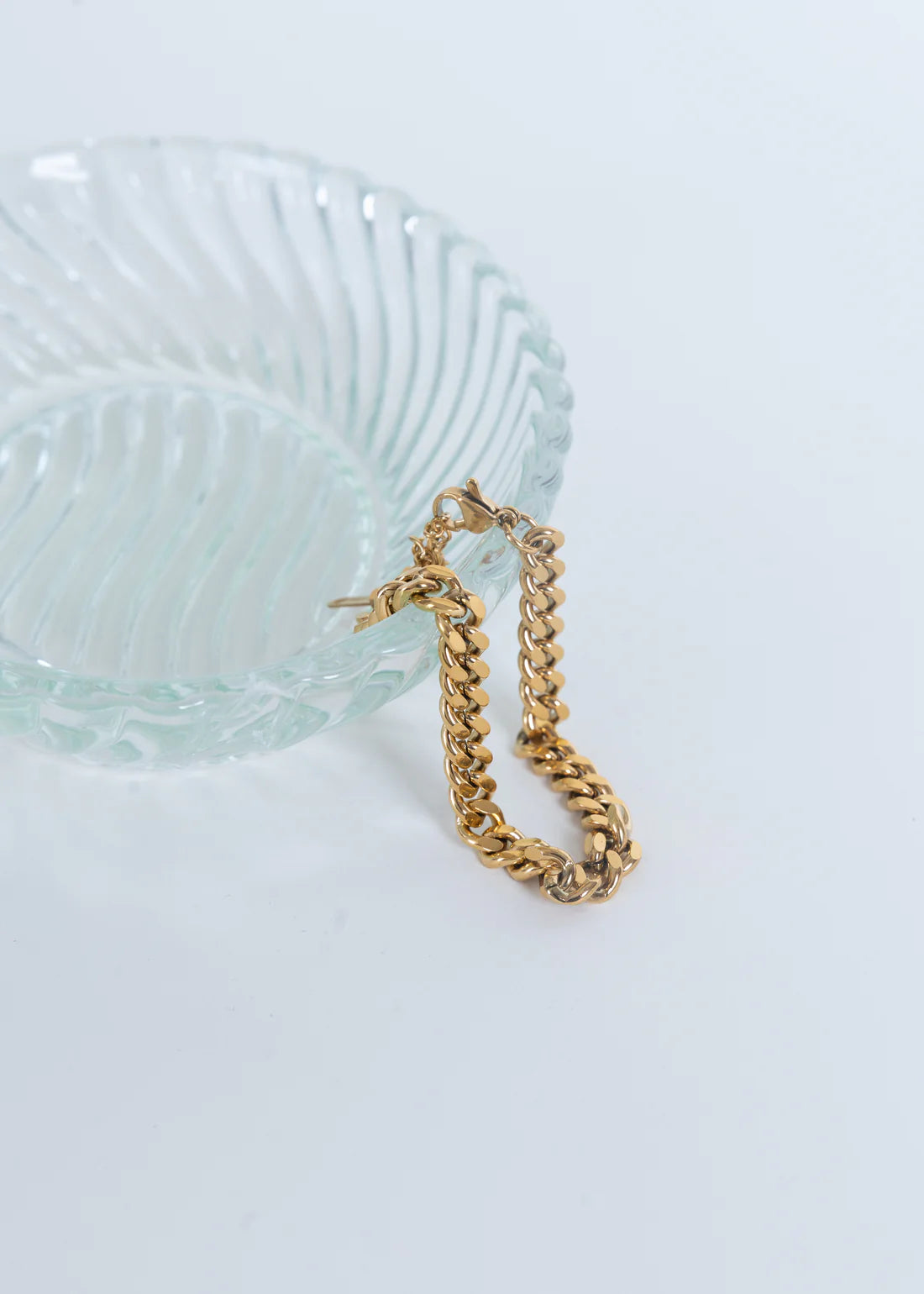 Gold chain bracelet draped over a clear glass bowl on a light blue background