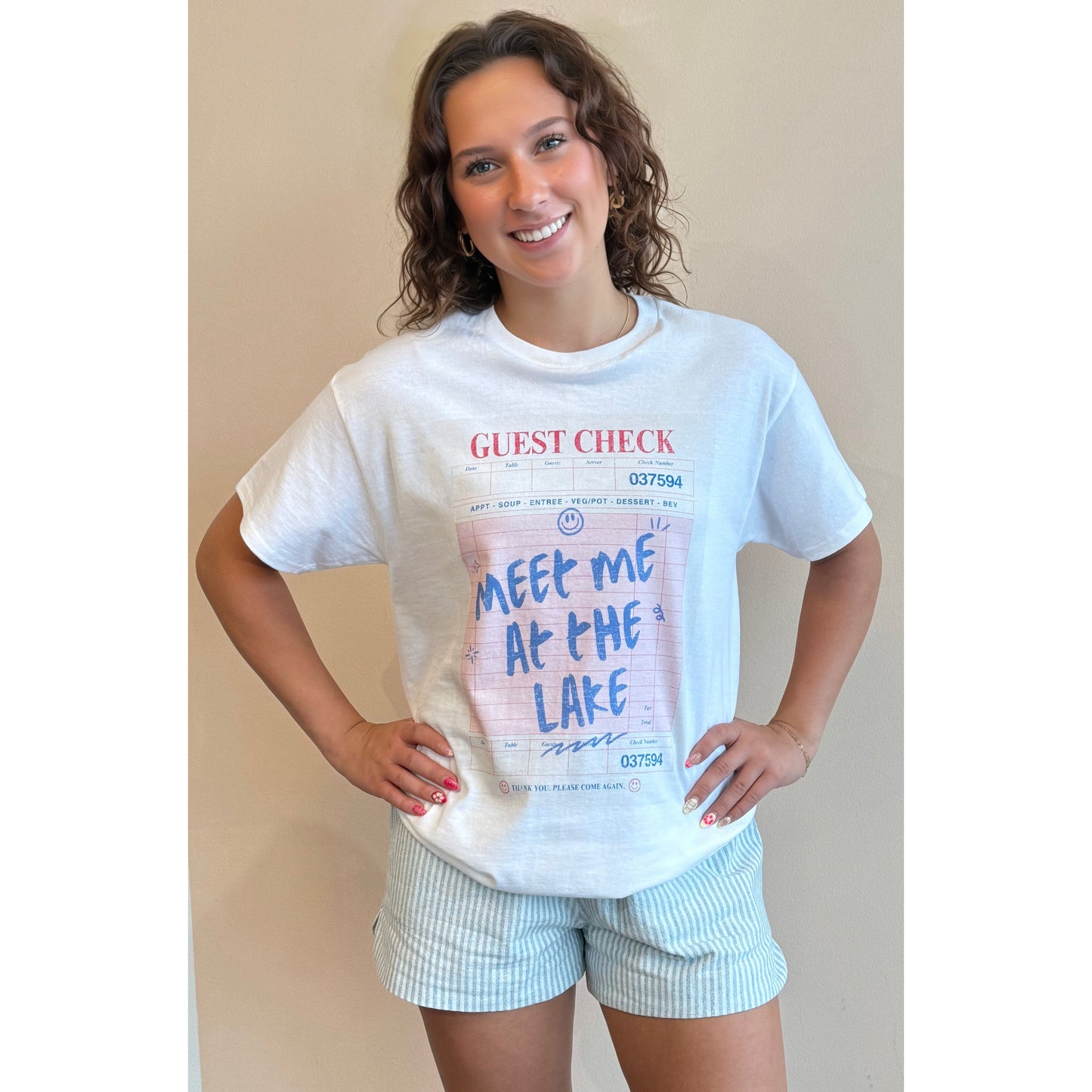 Woman wearing a white t-shirt with text and graphics, standing against a beige wall.
