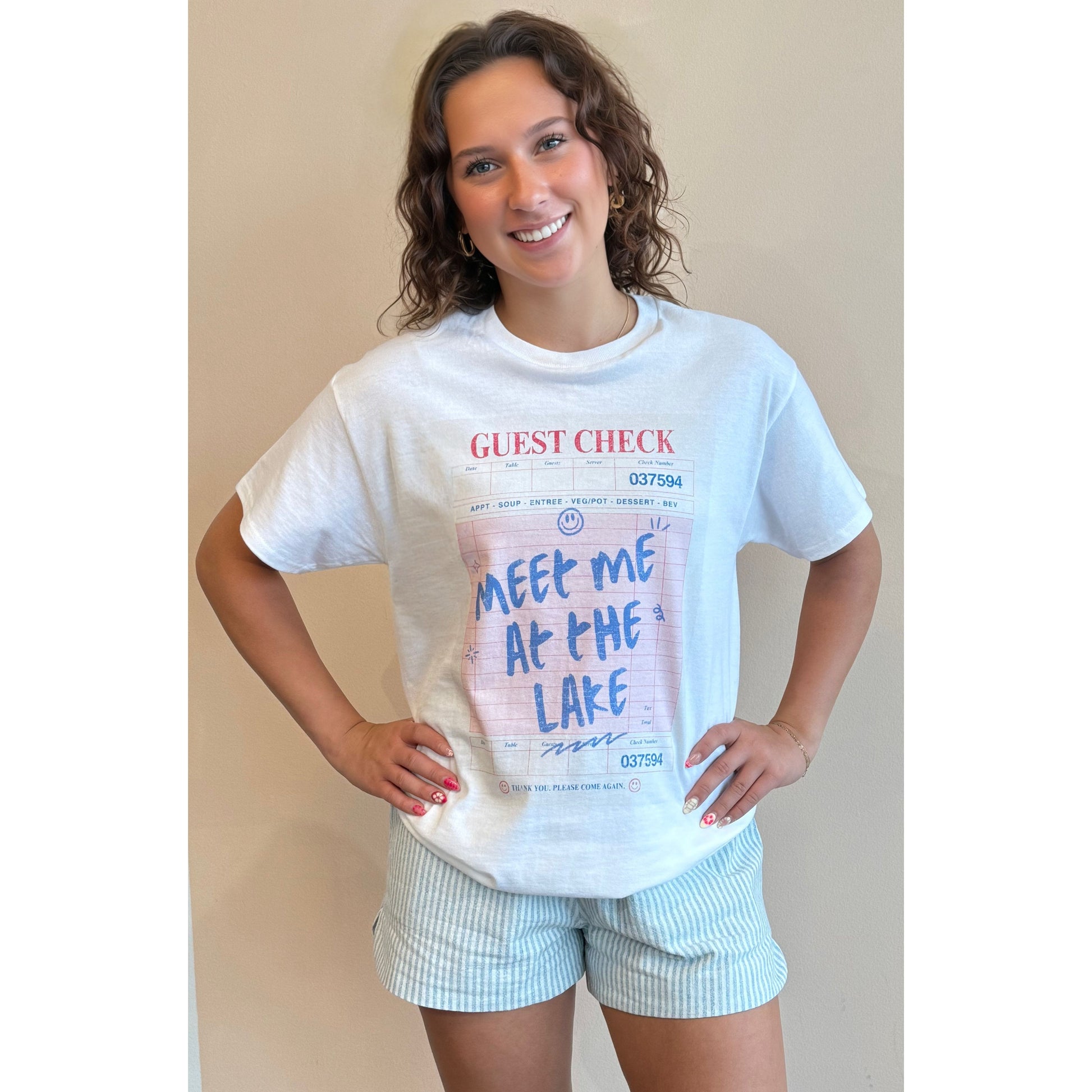 Woman wearing a white t-shirt with text and graphics, standing against a beige wall.