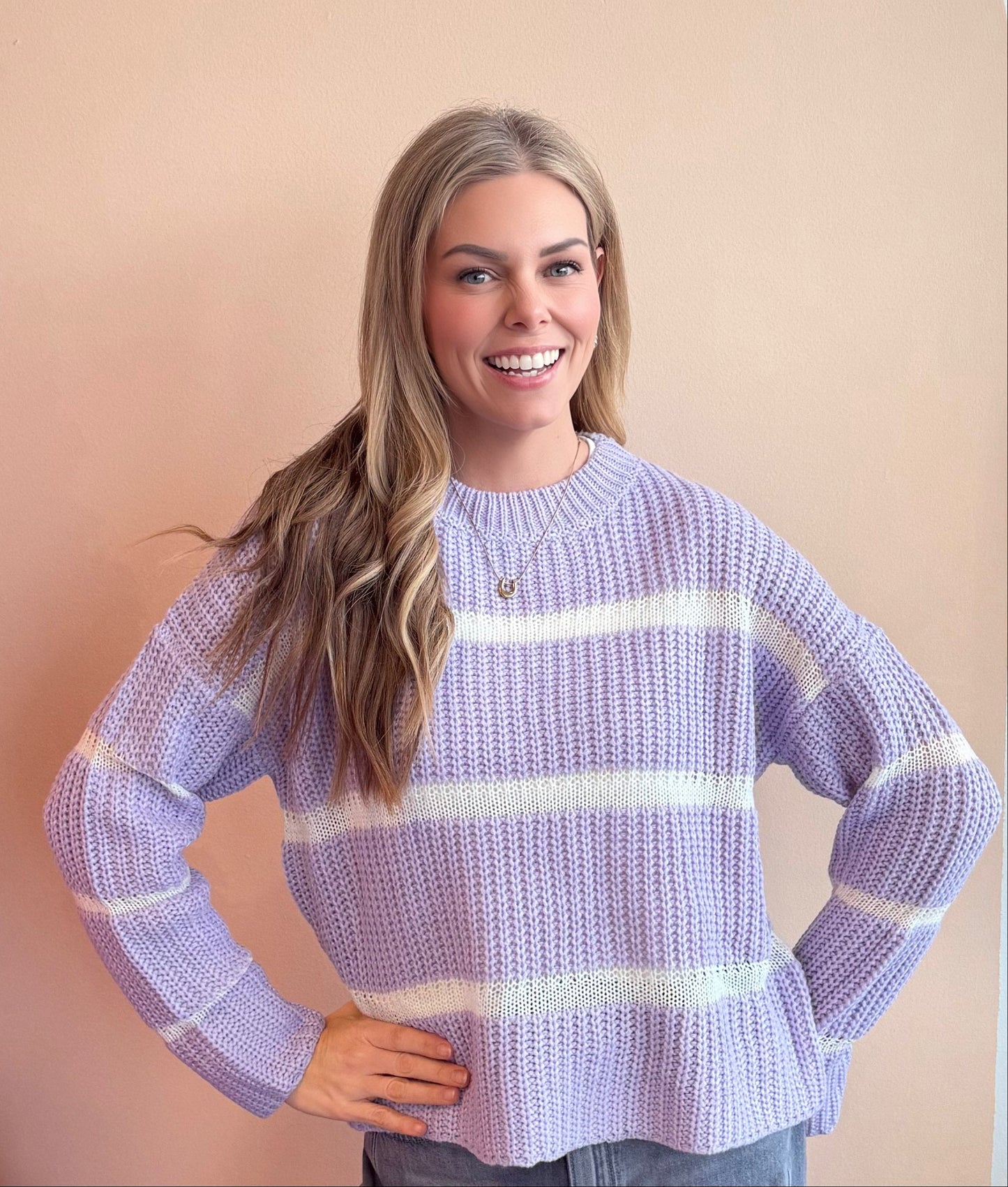 Woman wearing a light purple sweater with white stripes standing next to a pink Christmas tree against a beige background.