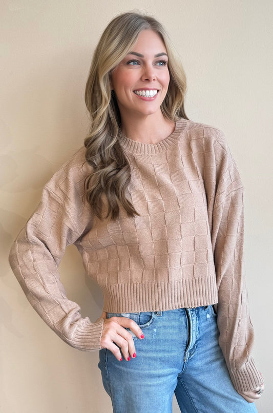 Woman wearing a checkboard pattern beige cropped sweater and blue jeans against a plain background