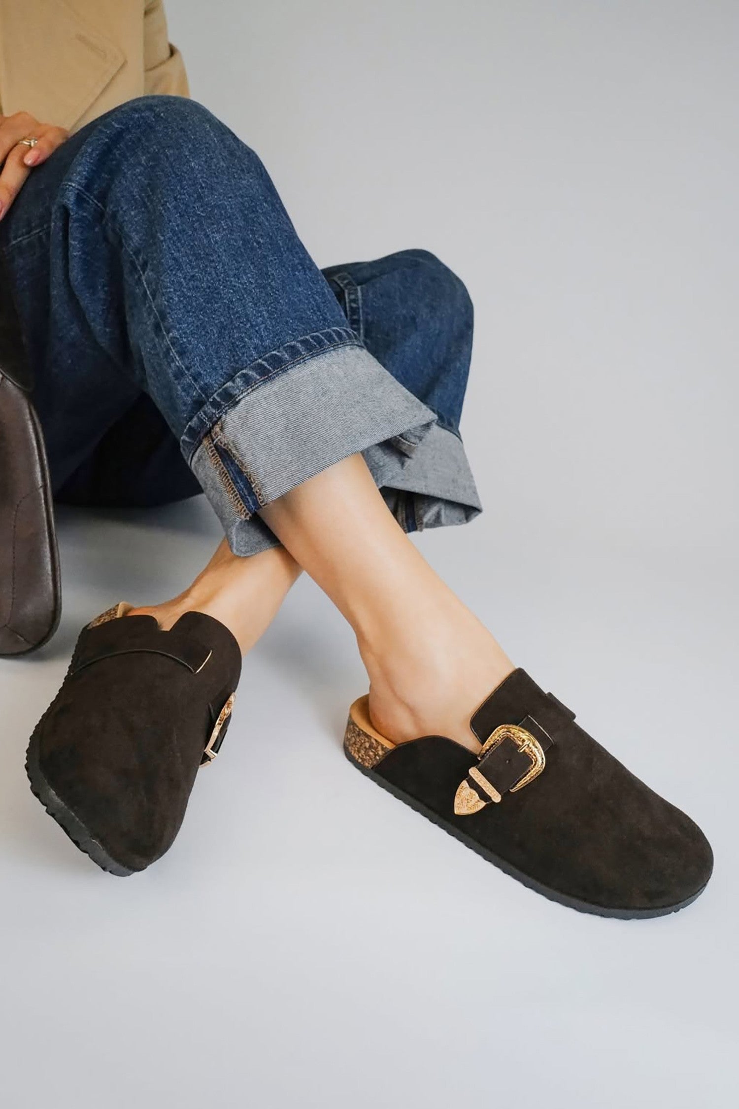 Person wearing black faux suede clogs with a gold buckle on a light gray background