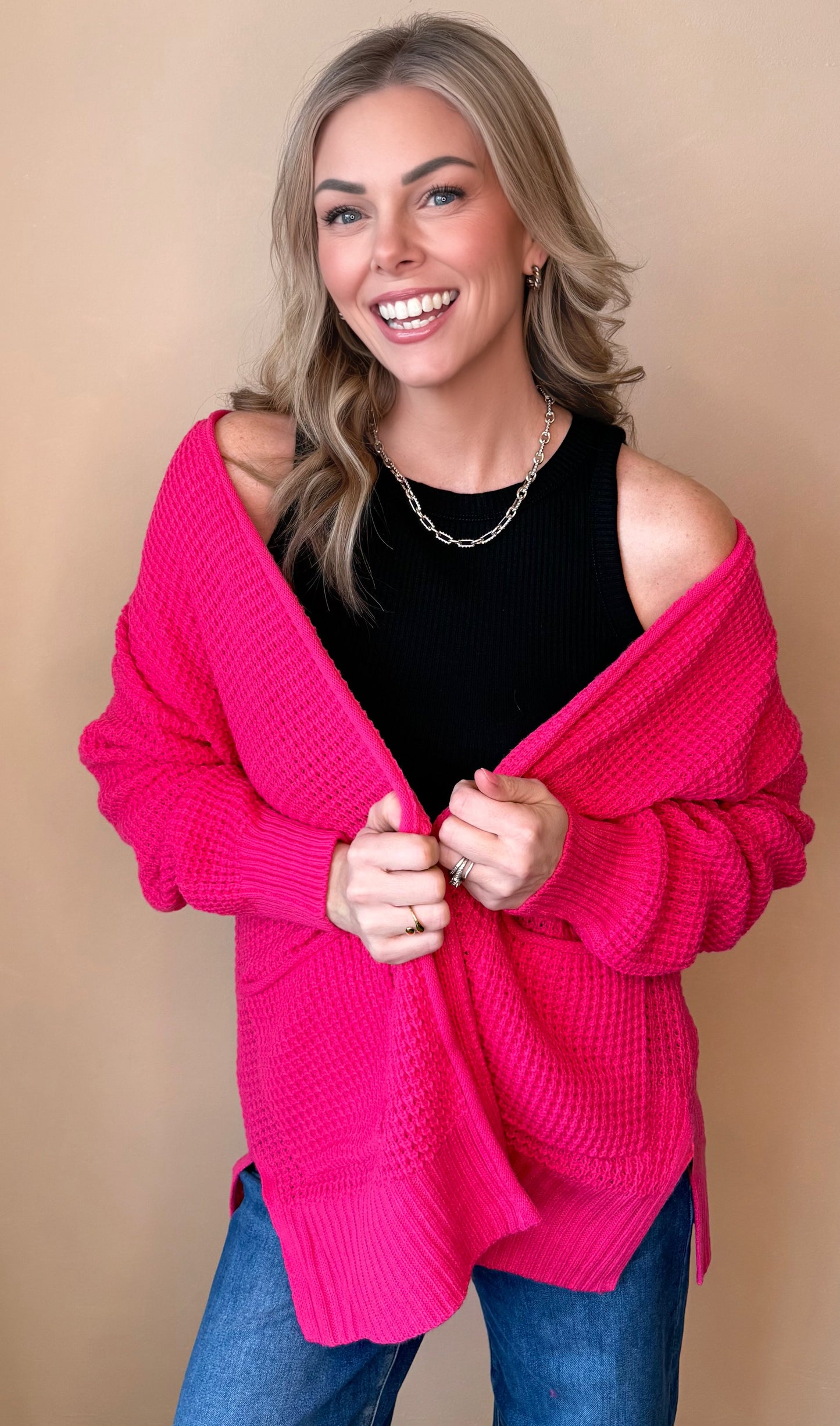 Woman wearing a bright pink cardigan over a black top and blue jeans on a beige background