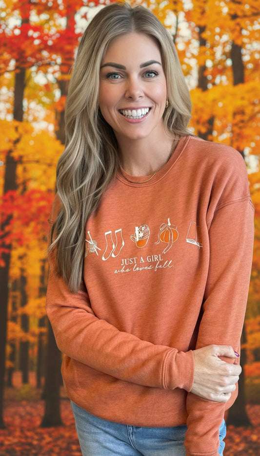Woman wearing an orange sweatshirt with a fall theme design screenprinted in beige outlines. Under fall graphics is a serif font in all caps says "Just a Girl" followed by a script font in lowercase "who loves fall"