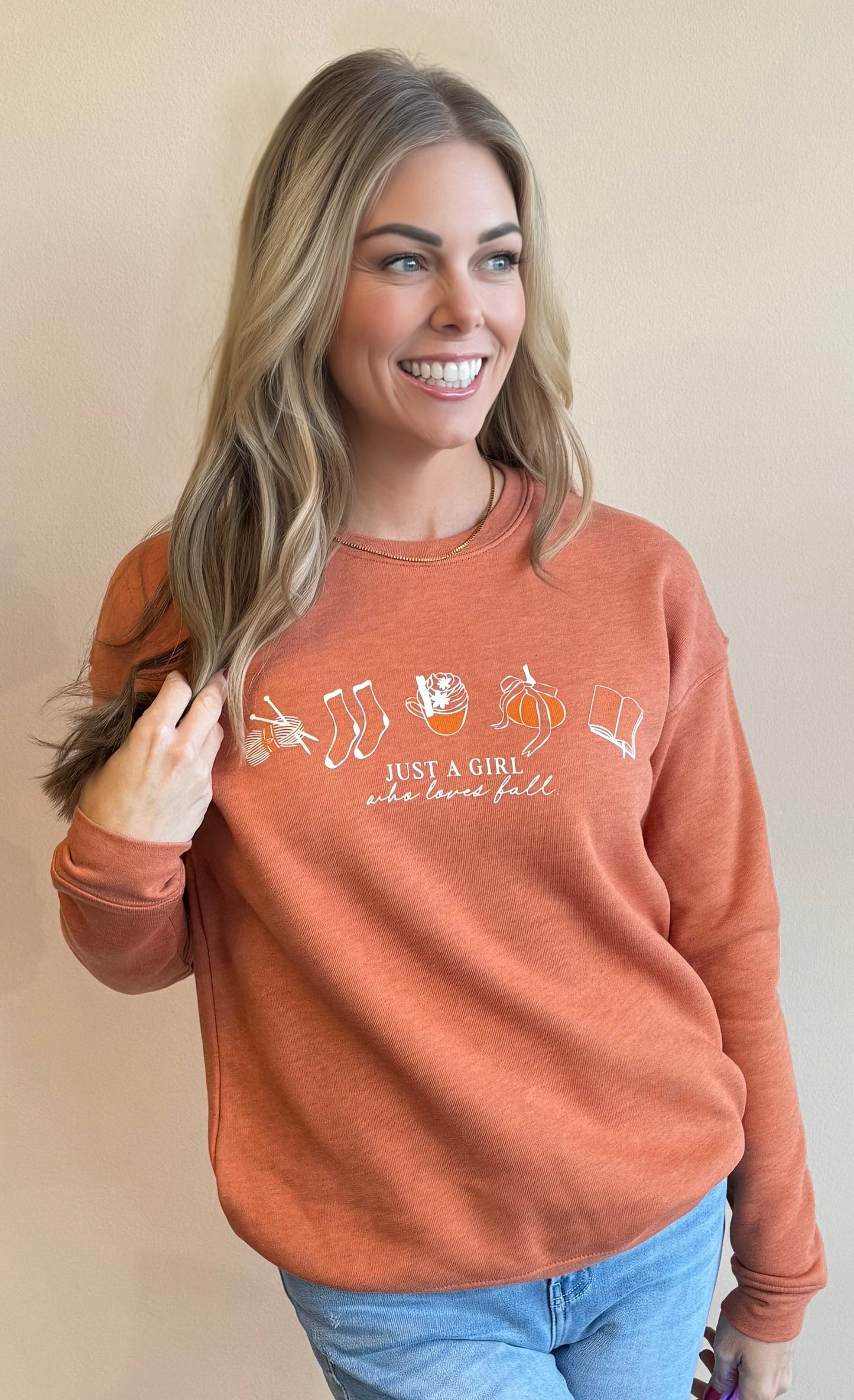 Woman wearing an orange sweatshirt with a fall theme design screenprinted in beige outlines. Under fall graphics is a serif font in all caps says "Just a Girl" followed by a script font in lowercase "who loves fall"