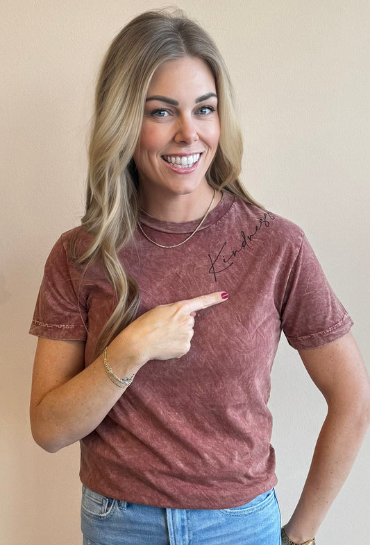 Woman smiling at the camera wearing a distressed maroon t-shirt pointing to the word "Kindness" in a script font screen printed on the left should of the shirt.