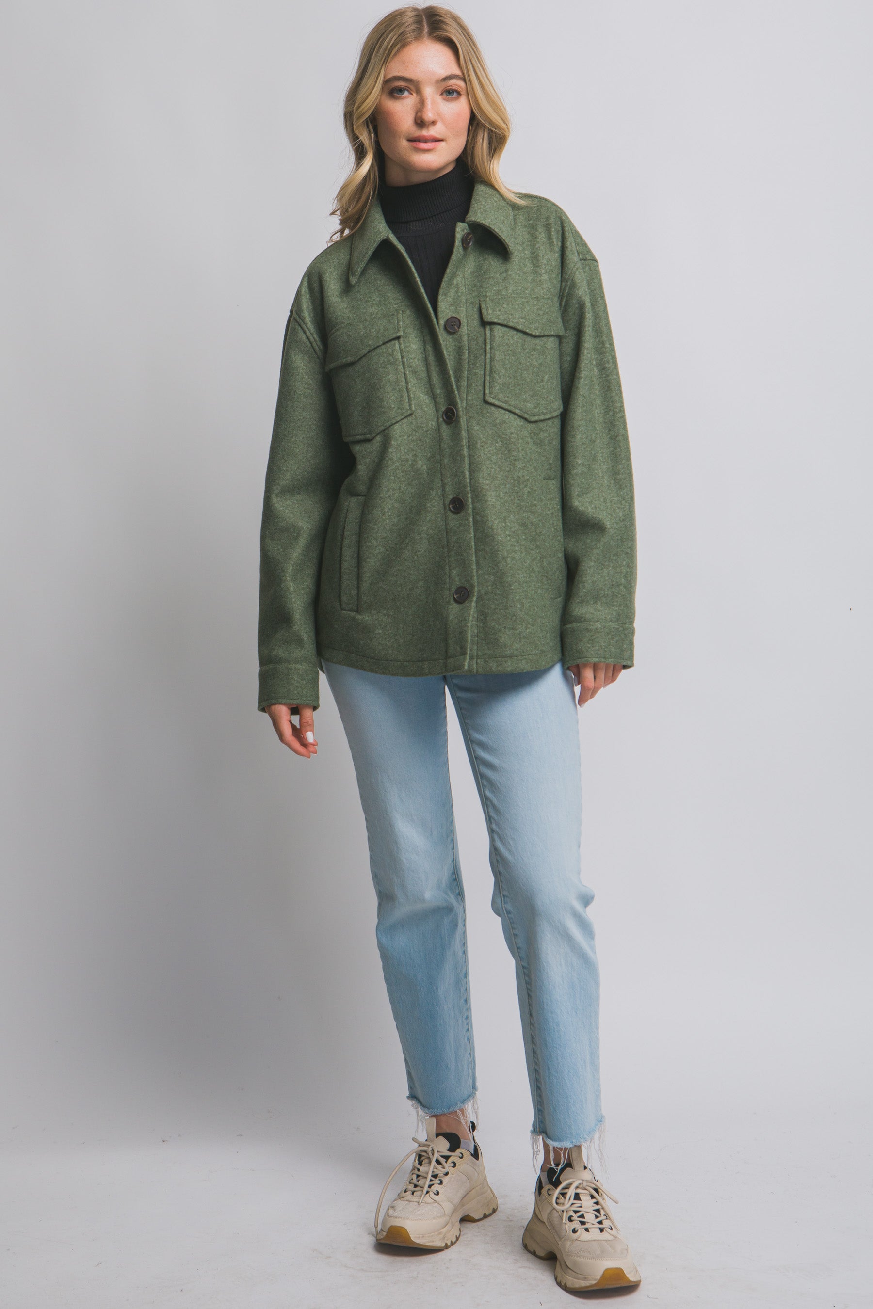 Woman wearing an olive green jacket, black turtleneck, light blue jeans, and beige sneakers on a gray background