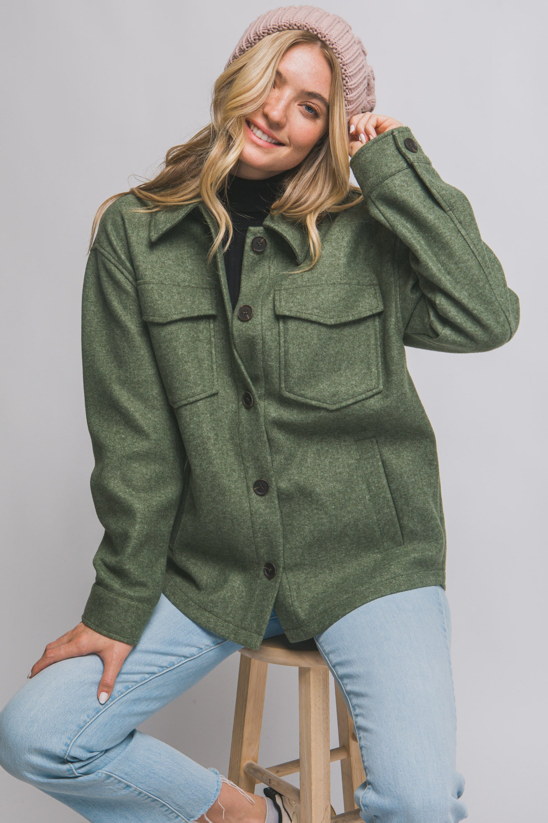 Woman wearing a green jacket and light blue jeans, sitting on a stool against a plain background