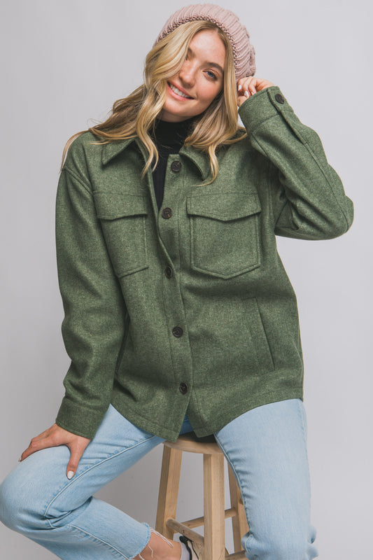 Woman wearing a green jacket and light blue jeans, sitting on a stool against a plain background
