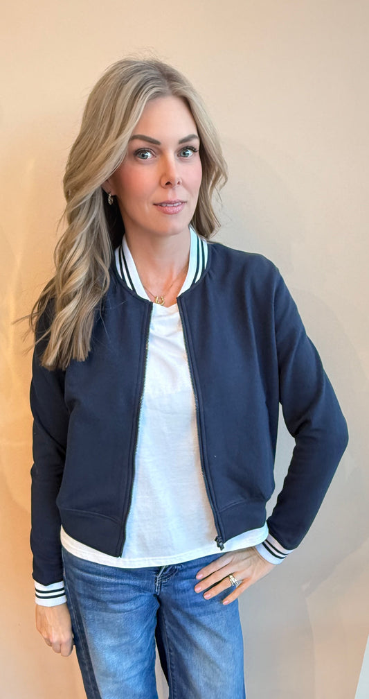 Woman wearing a navy bomber jacket, white shirt, and blue jeans indoors.