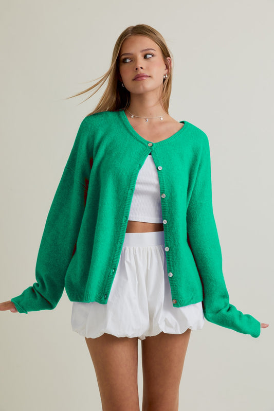 Woman wearing a clover green cardigan over a white top and shorts on a plain background