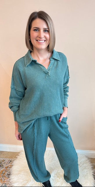Woman wearing a teal outfit standing in a room with a beige wall and plant.