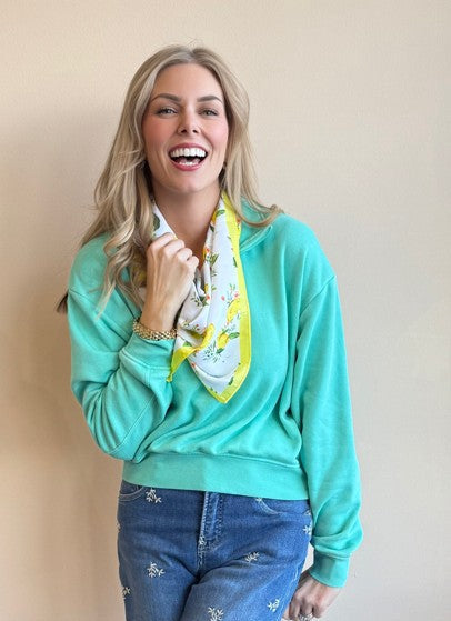 Woman wearing a teal sweatshirt and blue jeans with a yellow scarf, standing against a beige background.